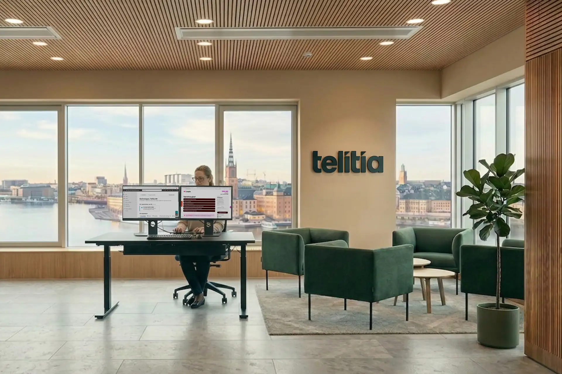En kvinna med glasögon sitter vid ett höj- och sänkbart skrivbord med två bildskärmar och arbetar, med en tydlig utsikt över Stockholm och Stadshuset genom ett stort fönster.